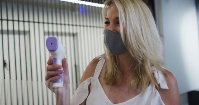 Woman is shown holding an infrared thermometer, likely conducting temperature checks in a healthcare setting or workplace to maintain safety measures during the Covid-19 pandemic. Useful for depicting concepts of safety, workplace health, and preventative measures against disease transmission.