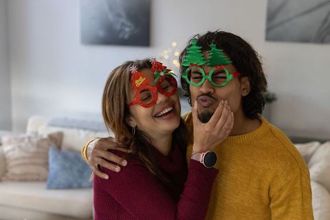 Couple wearing festive holiday glasses laughing and cuddling on cozy sofa, smartwatch visible