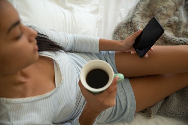 This image depicts a woman sitting comfortably at home, holding a cup of coffee and using her smartphone. Ideal for illustrating themes related to relaxation, morning routines, home lifestyle, quarantine, and technology use. Suitable for blogs, articles, and advertisements focusing on self-care, home living, and digital connectivity.