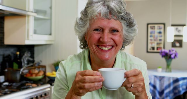 A senior Caucasian woman is smiling at the camera while holding a cup of coffee in a cozy kitchen setting, with copy space. Her cheerful expression and the domestic backdrop suggest a comfortable, homely atmosphere.