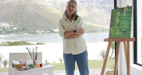 Mature woman artist standing with arms crossed by easel, green abstract canvas and seaside view