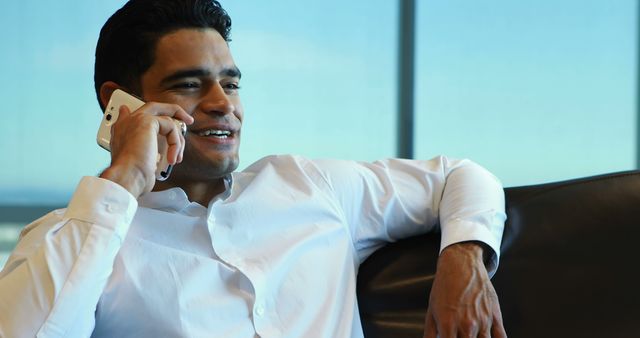 This image captures a young, confident businessman wearing a white shirt while talking on a smartphone in a modern office environment. Ideal for illustrative purposes depicting business communication, corporate settings, work culture, professional interactions, or advertisements related to office spaces, mobile communication services, and business consulting.