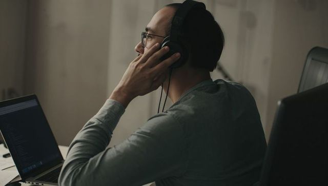 Focused developer wearing headphones coding on laptop in dim home office workspace