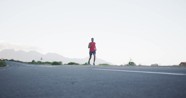 Man dressed in athletic gear running alone on an empty road with mountain background on early morning. Useful for illustrating concepts of fitness, endurance, outdoor recreation, healthy lifestyle, acquiring determination and toughness from morning exercise routines.