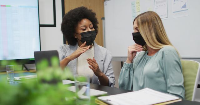 Businesswomen sitting at table discussing project while wearing face masks for safety. This image depicts a modern office environment during the covid-19 pandemic, highlighting collaboration, professionalism, and safety measures. Useful for illustrating concepts related to teamwork, business meetings, pandemic adaptation, and professional work settings.