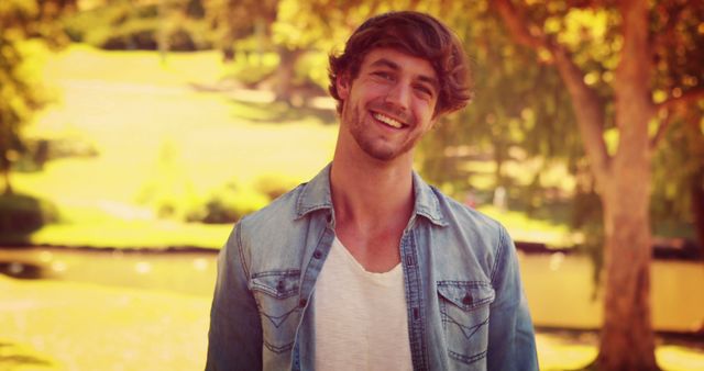 A young Caucasian man smiles warmly, standing outdoors in a sunny park setting, with copy space. His casual denim attire and relaxed demeanor suggest a laid-back, enjoyable day.