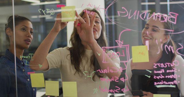 Three women collaborating on a project, using sticky notes and drawings on a transparent board. Ideal for depicting teamwork, creative brainstorming, and project planning in business settings. Perfect for articles on collaborative work methods, business strategy sessions, and innovative workspaces.