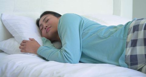 Mid-adult asian woman sleeping peacefully on white bedding wearing plaid pajama pants