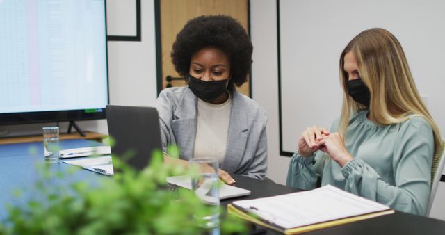 Two businesswomen sitting at a desk in a modern office, wearing masks and collaborating. They are working on a laptop and reviewing documents together. Suitable for use in articles or presentations about workplace health and safety, collaboration, diversity in business, or modern office environments.