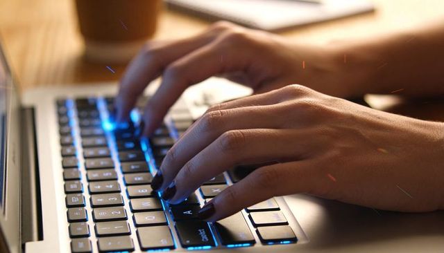 Close-up hands typing on backlit laptop keyboard on wooden desk with coffee notebook focus