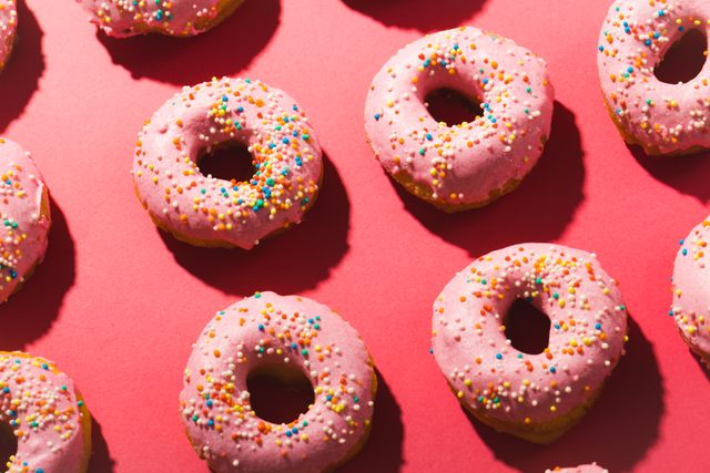This image shows a pattern of pink donuts with colorful sprinkles arranged on a pink background. Ideal for use in advertisements for bakeries, dessert shops, or food blogs. It can also be used in social media posts, marketing materials, or as a background for websites related to sweets and treats.