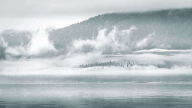 An ethereal scene captures mist rolling over layers of forested mountains, reflected on a calm lake. Perfect for conveying tranquility, serenity, and the untouched beauty of nature. Suitable for use in travel brochures, nature magazines, calming meditative spaces, and serene home decor.