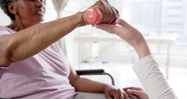 Elderly woman sitting in a wheelchair while using light weights for physical therapy. The presence of a healthcare professional providing assistance evidences the supportive care environment. This type of image is ideal for use in contexts related to elderly care, physical therapy advertisements, wellness programs, or senior fitness promotions, showcasing compassionate patient support and rehabilitation efforts.