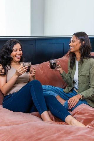 Two women laughing and sipping coffee on cozy rust sofa, chatting and relaxing indoors