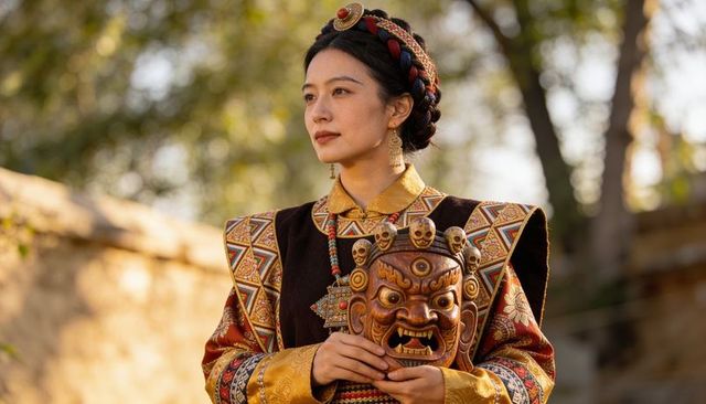 Woman holding carved ritual mask wearing ornate tibetan-style robe and braided crown