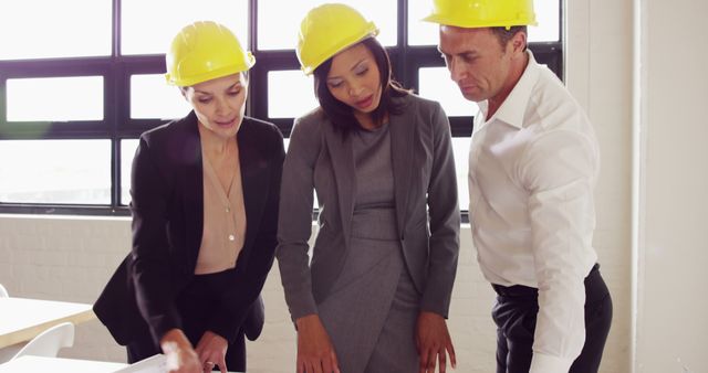 A diverse group of professionals, including a Caucasian woman and an African American woman, alongside a Caucasian man, all wearing hard hats, review plans together. They appear engaged in a serious discussion, related to construction or engineering, with copy space.