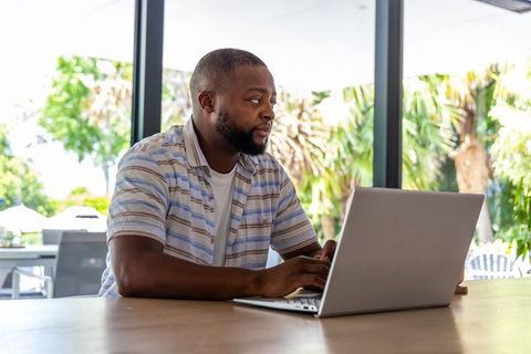 African american man working on laptop at modern wooden table with palm trees outside