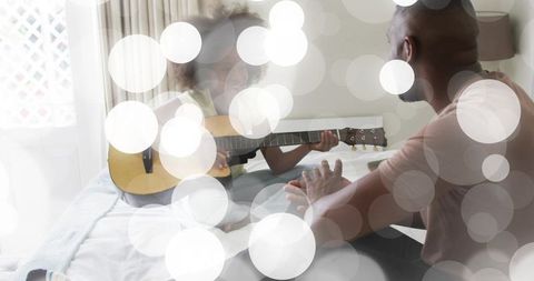 Young girl playing acoustic guitar on bed with parent during cozy home music session