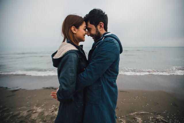 Couple standing close together and being affectionate on the beach with the ocean in background. They look happy and in love on an overcast day. Useful for marketing related to romantic getaways, relationship blogs, beach vacation packages or advertisements focused on love and connection.