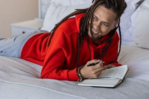 African american man lying on bed journaling and smiling while writing in notebook red hoodie