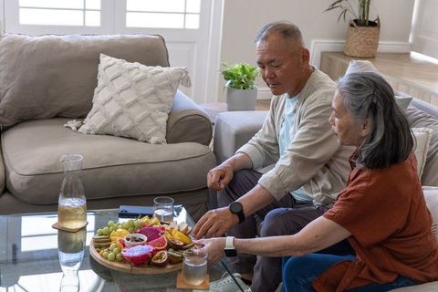 Asian senior couple sharing fruit snack board on cozy light-filled living room sofa