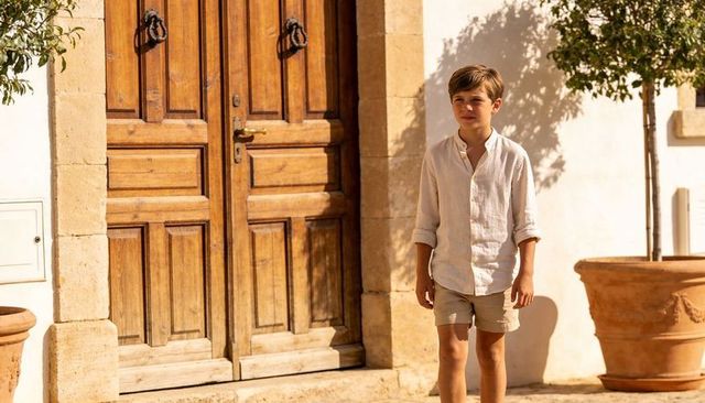 Young boy standing in sunlit mediterranean courtyard by rustic wooden door in linen shirt and shorts