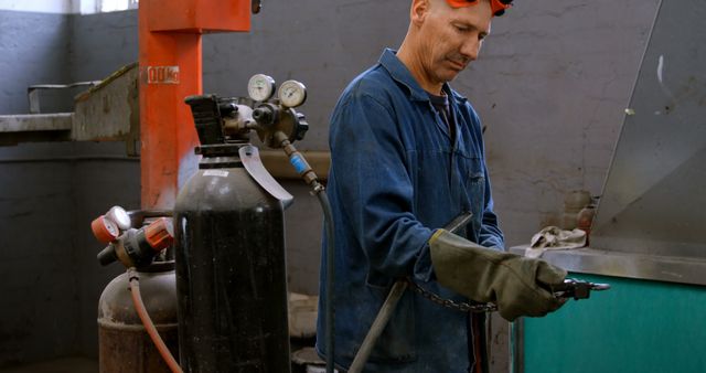 The image features a middle-aged man in a blue uniform and protective gloves handling welding equipment in an industrial facility. He appears focused and adept, emphasizing precision and safety in a manufacturing environment. This photo can be used for content related to industrial work, safety procedures, machinery, metalworking industries, skill development, and engineering fields, providing a realistic depiction of a skilled professional at work.