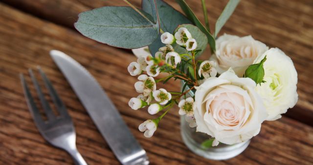 A delicate floral arrangement featuring pale roses and small white blossoms sits beside a fork and knife on a rustic wooden table, evoking a sense of elegance for a dining event. The composition suggests a carefully curated table setting for a special occasion or a fine dining experience.