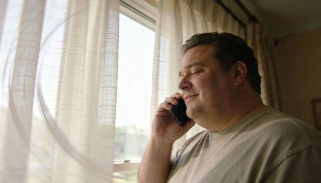 Middle-aged man talking on smartphone by sheer curtain window, relaxing in morning light