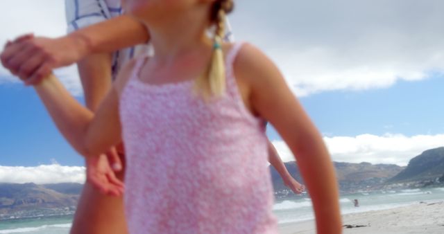A young girl in a pink dress is holding hands with an adult on a sunny beach, with copy space. Their bonding moment captures the carefree joy of a family vacation by the sea.