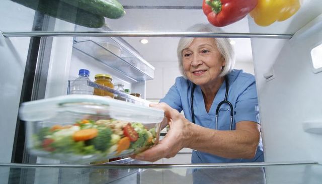 Senior nurse reaching for healthy salad in fridge wearing scrubs and stethoscope
