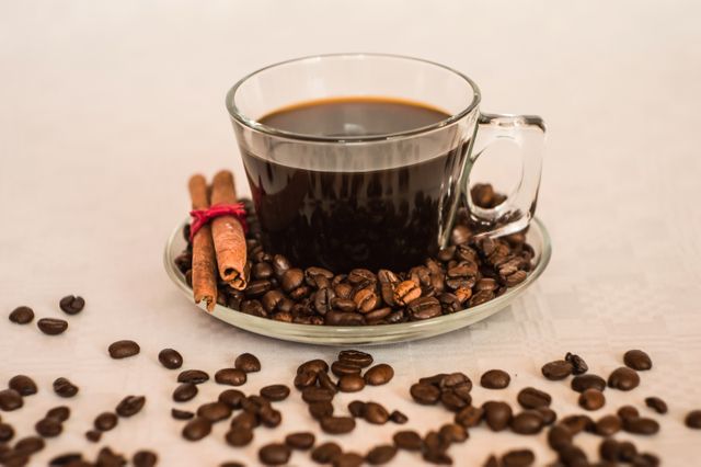 This image shows a glass mug filled with black coffee seated on a clear saucer. Scattered coffee beans encircle the mug and saucer, while a pair of cinnamon sticks is neatly tied together, adding a touch of spice to the warm and aromatic scene. This visual can be used for coffee-related advertisements, blogs focusing on morning routines or caffeine benefits, and promotional materials for cafes or coffee shops.