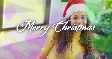Smiling woman wearing Santa hat decorating Christmas tree by window with festive glow