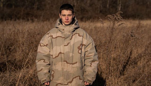 Young man standing wearing hooded desert camo parka in dry grass field at golden hour moody portrait