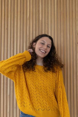 Smiling young woman wearing mustard knit sweater touching neck against wood slat wall