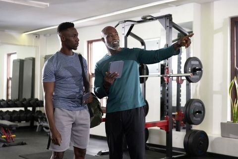 Personal trainer pointing while holding tablet coaching client near barbell rack in gym