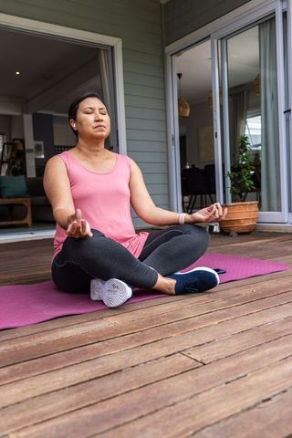 Mature woman practicing meditation on wooden deck with earbuds and yoga mat for mindfulness