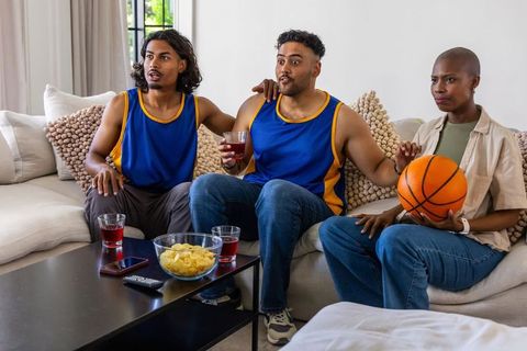 African american friends watching basketball game on couch holding basketball and snacks