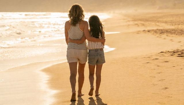 Walking mother and daughter arm in arm on golden beach at sunset, footprints in sand now