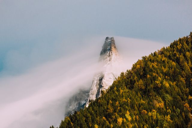 This image presents a dramatic mountain peak enveloped in mist, paired with the warm tones of a forest in autumn. Ideal for nature enthusiasts, travel blogs, outdoor adventure content, and environmental campaigns, it captures the beauty and serenity of untouched wilderness. It is perfect for uses that emphasize tranquility, natural beauty, and the allure of mysterious landscapes.