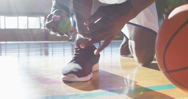 African american male basketball player tying shoes with team in background. basketball, sports training at an indoor court.