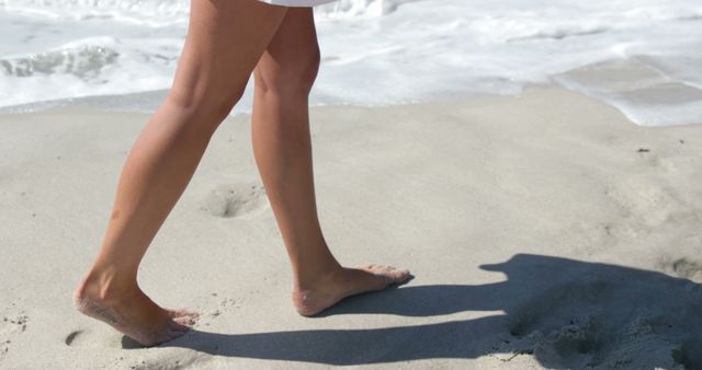 A close-up view of a woman's legs as she walks barefoot on a sandy beach, with gentle ocean waves lapping at the shore. This image conveys a sense of relaxation, summertime, and carefree moments. It can be used for travel brochures, vacation promotions, wellness articles, and lifestyle blogs.