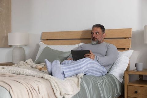 Mature man relaxing on bed in pajamas using tablet during cozy morning in neutral bedroom
