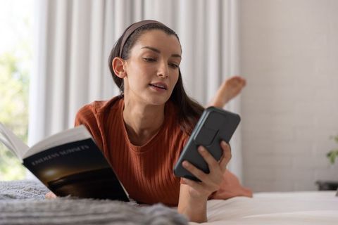 Mid-adult woman reading paperback while using smartphone on cozy minimalist bedroom bed