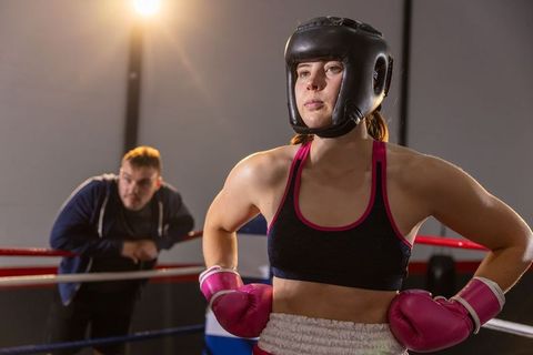 Female boxer preparing for fight under spotlight with coach leaning on ropes in ring