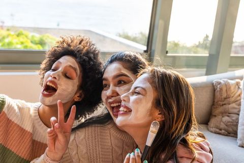 Three diverse friends laughing while enjoying at-home spa and wearing facial masks