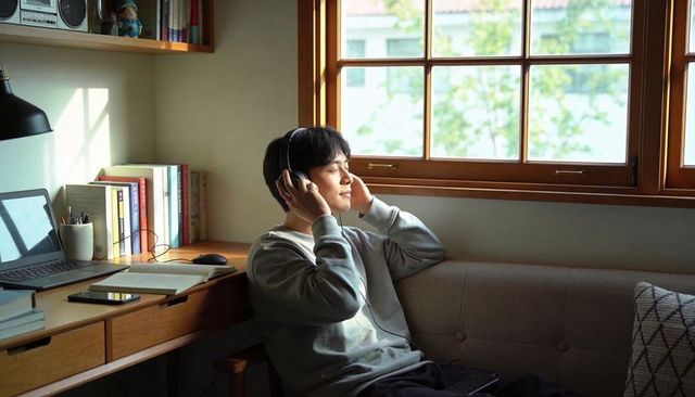 Young man relaxing and listening with over-ear headphones in home workspace by window