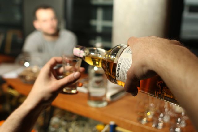Man's hand seen pouring whiskey into a glass at a bar counter. Blurred figure in the background creates a sense of social interaction. Perfect for use in contexts related to nightlife, social drinking, pubs, and relaxation.