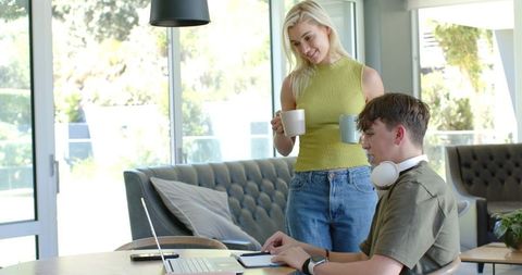 Parent offering coffee while teen studying with laptop and tablet in bright modern home