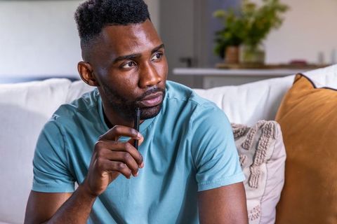 African american man sitting on sofa holding pen, contemplating at home, cozy lifestyle